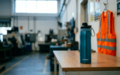 Garrafa térmica personalizada sobre mesa de fábrica com capacete de segurança e colete refletivo ao fundo